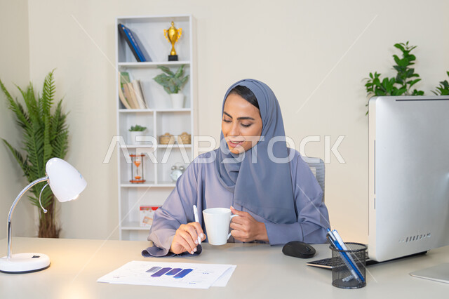 استخدام جهاز تقني حديث ومتطور، تحليل نتائج البيانات الاحصائية، امراة عربية خليجية اماراتية محجبة ترتدي العباءة تجلس خلف الطاولة وبجانبها مجموعة مخططات تعمل على الحاسب لانجاز المهام في المكتب، سيدة سعودية تدون الملاحظات على الورق، وظيفة مكتبية في شركات الخليج العربي
