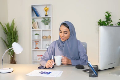 استخدام جهاز تقني حديث ومتطور، تحليل نتائج البيانات الاحصائية، امراة عربية خليجية اماراتية محجبة ترتدي العباءة تجلس خلف الطاولة وبجانبها مجموعة مخططات تعمل على الحاسب لانجاز المهام في المكتب، سيدة سعودية تدون الملاحظات على الورق، وظيفة مكتبية في شركات الخليج العربي