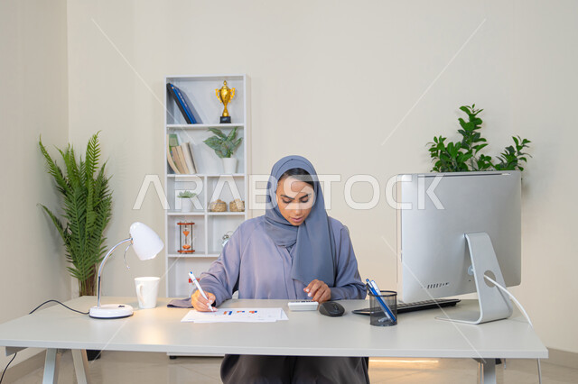 سيدة سعودية تدون الملاحظات على الورق، استخدام الالة الحاسبة لاجراء العمليات الحسابية، وظيفة مكتبية في شركات الخليج العربي، تحليل نتائج البيانات الاحصائية، امراة عربية خليجية اماراتية محجبة ترتدي العباءة تجلس خلف الطاولة تعمل على الحاسب لانجاز المهام في المكتب