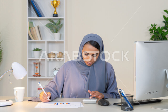 تحليل نتائج البيانات الاحصائية، امراة عربية خليجية اماراتية محجبة ترتدي العباءة تجلس خلف الطاولة تعمل على الحاسب لانجاز المهام في المكتب، سيدة سعودية تدون الملاحظات على الورق، استخدام الالة الحاسبة لاجراء العمليات الحسابية، وظيفة مكتبية في شركات الخليج العربي