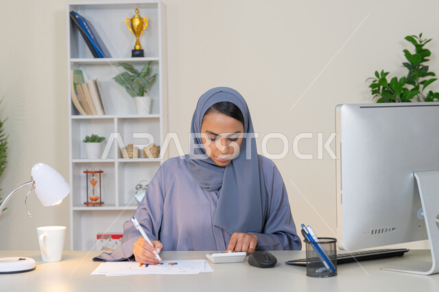 سيدة سعودية تدون الملاحظات على الورق، استخدام الالة الحاسبة لاجراء العمليات الحسابية، وظيفة مكتبية في شركات الخليج العربي، تحليل نتائج البيانات الاحصائية، امراة عربية خليجية اماراتية محجبة ترتدي العباءة تجلس خلف الطاولة تعمل على الحاسب لانجاز المهام في المكتب