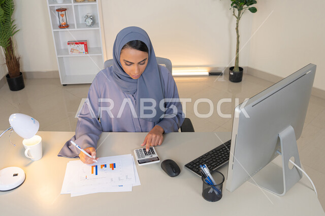A veiled Emirati Gulf Arab woman wearing an abaya sitting behind a desk working on a computer to complete tasks in the office, a Saudi woman taking notes on paper, using a calculator to perform calculations, an office job in a Gulf company, analyzing statistical data results