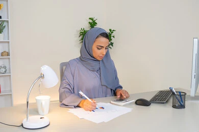 وظيفة مكتبية في شركات الخليج العربي، تحليل نتائج البيانات الاحصائية، امراة عربية خليجية اماراتية محجبة ترتدي العباءة تجلس خلف الطاولة تعمل على الحاسب لانجاز المهام في المكتب، سيدة سعودية تدون الملاحظات على الورق، استخدام الالة الحاسبة لاجراء العمليات الحسابية