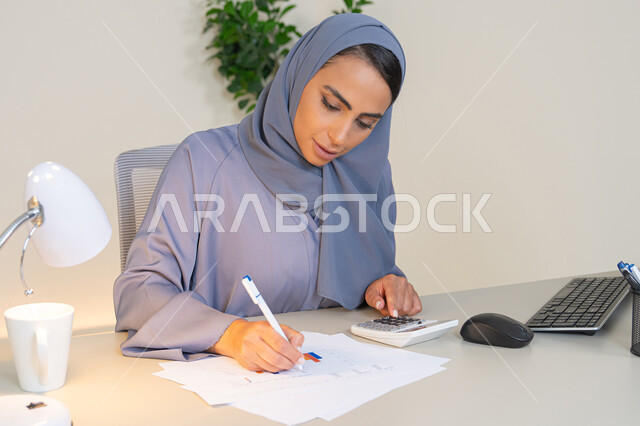 امراة عربية خليجية اماراتية محجبة ترتدي العباءة تجلس خلف الطاولة تعمل على الحاسب لانجاز المهام في المكتب، سيدة سعودية تدون الملاحظات على الورق، استخدام الالة الحاسبة لاجراء العمليات الحسابية، وظيفة مكتبية في شركات الخليج العربي، تحليل نتائج البيانات الاحصائية