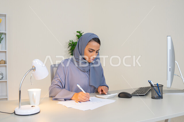 تحليل نتائج البيانات الاحصائية، امراة عربية خليجية اماراتية محجبة ترتدي العباءة تجلس خلف الطاولة تعمل على الحاسب لانجاز المهام في المكتب، سيدة سعودية تدون الملاحظات على الورق، استخدام الالة الحاسبة لاجراء العمليات الحسابية، وظيفة مكتبية في شركات الخليج العربي
