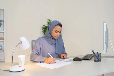 تحليل نتائج البيانات الاحصائية، امراة عربية خليجية اماراتية محجبة ترتدي العباءة تجلس خلف الطاولة تعمل على الحاسب لانجاز المهام في المكتب، سيدة سعودية تدون الملاحظات على الورق، استخدام الالة الحاسبة لاجراء العمليات الحسابية، وظيفة مكتبية في شركات الخليج العربي