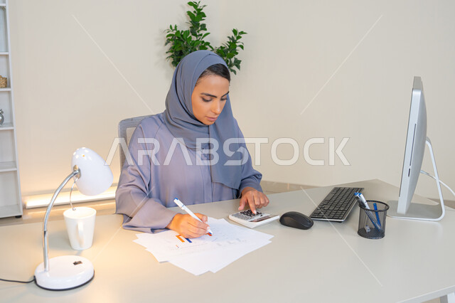 Analysis of statistical data results, a veiled Emirati Gulf Arab woman wearing an abaya sitting behind a desk working on a computer to complete tasks in the office, a Saudi woman taking notes on paper, using a calculator to perform mathematical operations, an office job in a Gulf Arab company