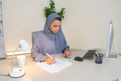 تحليل نتائج البيانات الاحصائية، امراة عربية خليجية اماراتية محجبة ترتدي العباءة تجلس خلف الطاولة تعمل على الحاسب لانجاز المهام في المكتب، سيدة سعودية تدون الملاحظات على الورق، استخدام الالة الحاسبة لاجراء العمليات الحسابية، وظيفة مكتبية في شركات الخليج العربي