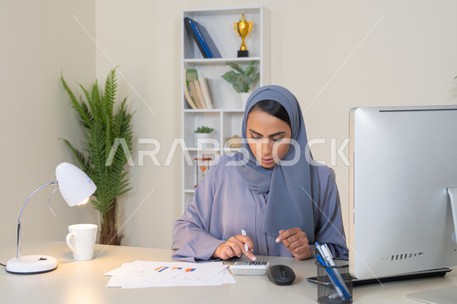 وظيفة مكتبية في شركات الخليج العربي، تحليل نتائج البيانات الاحصائية، امراة عربية خليجية اماراتية محجبة ترتدي العباءة تجلس خلف الطاولة تعمل على الحاسب لانجاز المهام في المكتب، سيدة سعودية تدون الملاحظات على الورق، استخدام الالة الحاسبة لاجراء العمليات الحسابية