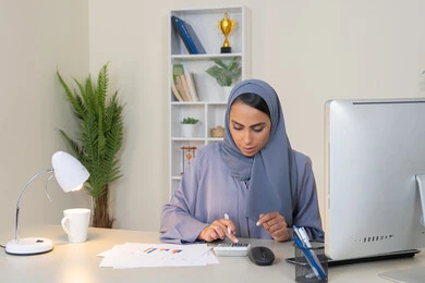 Office job in a Gulf company, analyzing statistical data results, a veiled Emirati Gulf Arab woman wearing an abaya sitting behind a desk working on a computer to complete tasks in the office, a Saudi woman taking notes on paper, using a calculator to perform mathematical operations