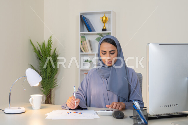 امراة عربية خليجية اماراتية محجبة ترتدي العباءة تجلس خلف الطاولة تعمل على الحاسب لانجاز المهام في المكتب، سيدة سعودية تدون الملاحظات على الورق، استخدام الالة الحاسبة لاجراء العمليات الحسابية، وظيفة مكتبية في شركات الخليج العربي، تحليل نتائج البيانات الاحصائية