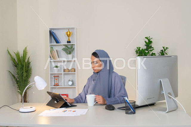 ادارة الأعمال وانجاز المهام المطلوبة، العمل عن بعد، مهنة ووظيفة نسائية، إمرأة عربية خليجية اماراتية محجبة ترتدي عباءة ملونة تجلس خلف الطاولة تستخدم الجهاز اللوحي وتشرب القهوة، سيدة سعودية تستخدم جهاز تقني حديث ومتطور في المكتب، احتساء المشروب المفضل في المكتب