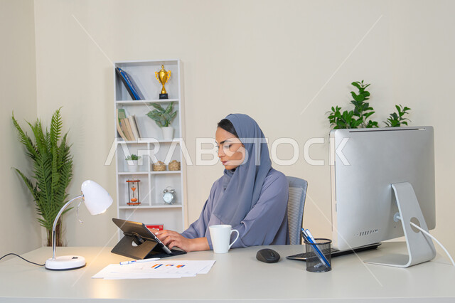 Remote work, female profession and job, veiled Emirati Gulf Arab woman wearing a colorful abaya sitting behind a table using a tablet and drinking coffee, Saudi woman using a modern and advanced technical device in the office, drinking her favorite drink in the office, business management and completing required tasks