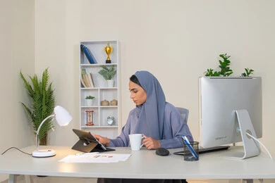 Remote work, female profession and job, veiled Emirati Gulf Arab woman wearing a colorful abaya sitting behind a table using a tablet and drinking coffee, Saudi woman using a modern and advanced technical device in the office, drinking her favorite drink in the office, business management and completing required tasks