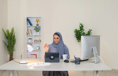 Raising the palm of the hand with a greeting gesture, a female profession and job, a veiled Emirati Gulf Arab woman wearing a colorful abaya sitting behind a table using a tablet and drinking coffee, a Saudi woman making a video call in the office, business management and completing required tasks, working remotely