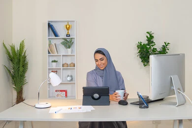 Business administration and completing required tasks, remote work, female profession and job, veiled Emirati Gulf Arab woman wearing a colorful abaya sitting behind a table using a tablet and drinking coffee, Saudi woman using a modern and advanced technical device in the office, drinking her favorite drink in the office