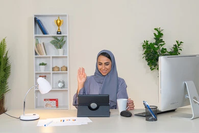 مهنة ووظيفة نسائية، إمرأة عربية خليجية اماراتية محجبة ترتدي عباءة ملونة تجلس خلف الطاولة تستخدم الجهاز اللوحي وتشرب القهوة، سيدة سعودية تجري مكالمة فيديو في المكتب، ادارة الأعمال وانجاز المهام المطلوبة، العمل عن بعد، رفع كف اليد بايماءات القاء التحية والسلام