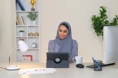 Business administration and completing required tasks, remote work, female profession and job, veiled Emirati Gulf Arab woman wearing a colorful abaya sitting behind a table using a tablet and drinking coffee, Saudi woman using a modern and advanced technical device in the office, drinking her favorite drink in the office