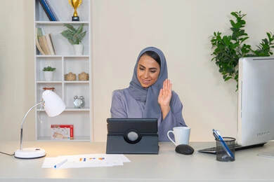 رفع كف اليد بايماءات القاء التحية والسلام، مهنة ووظيفة نسائية، إمرأة عربية خليجية اماراتية محجبة ترتدي عباءة ملونة تجلس خلف الطاولة تستخدم الجهاز اللوحي وتشرب القهوة، سيدة سعودية تجري مكالمة فيديو في المكتب، ادارة الأعمال وانجاز المهام المطلوبة، العمل عن بعد
