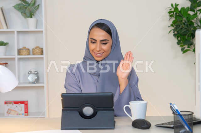 رفع كف اليد بايماءات القاء التحية والسلام، مهنة ووظيفة نسائية، إمرأة عربية خليجية اماراتية محجبة ترتدي عباءة ملونة تجلس خلف الطاولة تستخدم الجهاز اللوحي وتشرب القهوة، سيدة سعودية تجري مكالمة فيديو في المكتب، ادارة الأعمال وانجاز المهام المطلوبة، العمل عن بعد