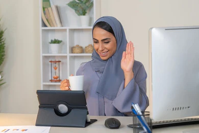 A female profession and job, a veiled Emirati Gulf Arab woman wearing a colorful abaya sitting behind a table using a tablet and drinking coffee, a Saudi woman making a video call in the office, managing business and completing required tasks, working remotely, raising her hand with gestures of greeting and peace