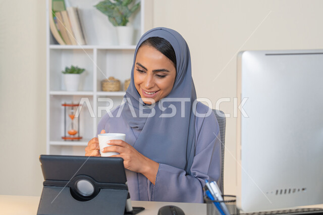 Business administration and completing required tasks, remote work, female profession and job, veiled Emirati Gulf Arab woman wearing a colorful abaya sitting behind a table using a tablet and drinking coffee, Saudi woman using a modern and advanced technical device in the office, drinking her favorite drink in the office