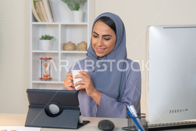 A female profession and job, a veiled Emirati Gulf Arab woman wearing a colorful abaya sitting behind a table using a tablet and drinking coffee, a Saudi woman using a modern and advanced technical device in the office, drinking her favorite drink in the office, managing business and completing required tasks, working remotely