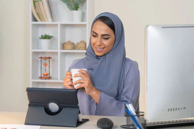 A female profession and job, a veiled Emirati Gulf Arab woman wearing a colorful abaya sitting behind a table using a tablet and drinking coffee, a Saudi woman using a modern and advanced technical device in the office, drinking her favorite drink in the office, managing business and completing required tasks, working remotely
