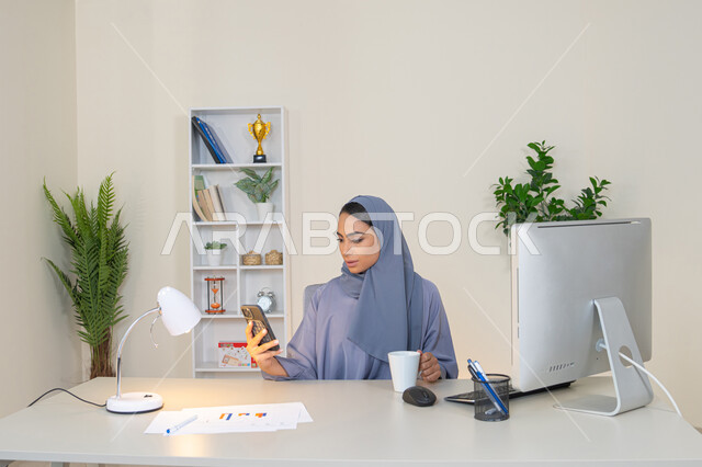 احتساء المشروب المفضل في المكتب، سيدة سعودية تتصفح التطبيقات وبرامج التواصل الاجتماعي، تبادل الأحاديث والأخبار مع الأهل والأصدقاء،  امراة عربية خليجية اماراتية ترتدي العباءة والحجاب تمسك بيدها الهاتف المحمول وتشرب القهوة، استخدام جهاز تقني حديث ومتطور