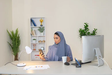 احتساء المشروب المفضل في المكتب، سيدة سعودية تتصفح التطبيقات وبرامج التواصل الاجتماعي، تبادل الأحاديث والأخبار مع الأهل والأصدقاء،  امراة عربية خليجية اماراتية ترتدي العباءة والحجاب تمسك بيدها الهاتف المحمول وتشرب القهوة، استخدام جهاز تقني حديث ومتطور