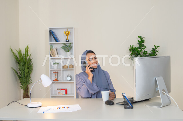 Using a modern and advanced technical device, a Saudi woman sitting behind a table and drinking her favorite drink in the office, making a phone call via a mobile device, exchanging conversations and news and communicating with family and friends, an Emirati woman wearing an abaya and hijab holding a mobile phone in her hand and drinking coffee