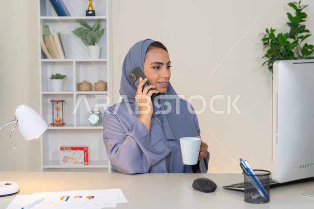 Exchanging conversations and news and communicating with family and friends, an Emirati woman wearing an abaya and hijab holding a mobile phone and drinking coffee, using a modern and advanced technical device, a Saudi woman sitting behind a table and sipping her favorite drink in the office, making a phone call via a mobile device