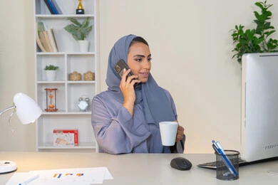 Exchanging conversations and news and communicating with family and friends, an Emirati woman wearing an abaya and hijab holding a mobile phone and drinking coffee, using a modern and advanced technical device, a Saudi woman sitting behind a table and sipping her favorite drink in the office, making a phone call via a mobile device