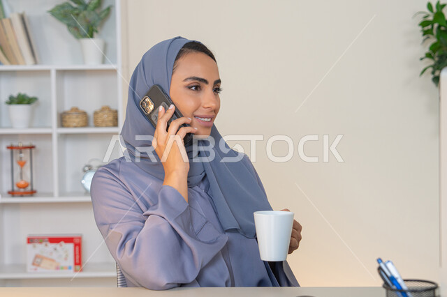 التواصل وتبادل الأحاديث والأخبار مع الأهل والأصدقاء،  امراة اماراتية ترتدي العباءة والحجاب تمسك بيدها الهاتف المحمول وتشرب القهوة، استخدام جهاز تقني حديث ومتطور، سيدة سعودية تجلس خلف الطاولة وتحتسي المشروب المفضل في المكتب، اجراء مكالمة هاتفية عبر جهاز الجوال
