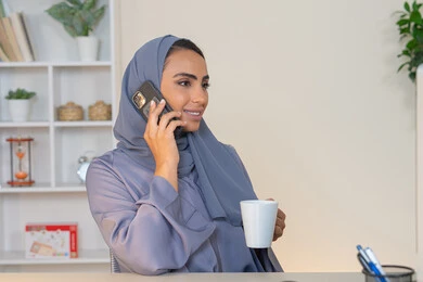 التواصل وتبادل الأحاديث والأخبار مع الأهل والأصدقاء،  امراة اماراتية ترتدي العباءة والحجاب تمسك بيدها الهاتف المحمول وتشرب القهوة، استخدام جهاز تقني حديث ومتطور، سيدة سعودية تجلس خلف الطاولة وتحتسي المشروب المفضل في المكتب، اجراء مكالمة هاتفية عبر جهاز الجوال
