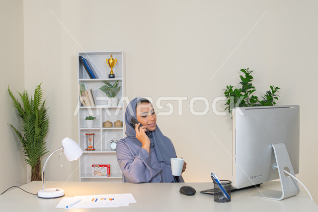اجراء مكالمة هاتفية عبر جهاز الجوال، التواصل وتبادل الأحاديث والأخبار مع الأهل والأصدقاء،  امراة اماراتية ترتدي العباءة والحجاب تمسك بيدها الهاتف المحمول وتشرب القهوة، استخدام جهاز تقني حديث ومتطور، سيدة سعودية تجلس خلف الطاولة وتحتسي المشروب المفضل في المكتب