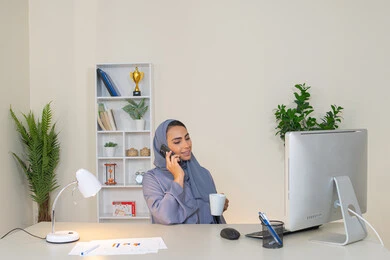 اجراء مكالمة هاتفية عبر جهاز الجوال، التواصل وتبادل الأحاديث والأخبار مع الأهل والأصدقاء،  امراة اماراتية ترتدي العباءة والحجاب تمسك بيدها الهاتف المحمول وتشرب القهوة، استخدام جهاز تقني حديث ومتطور، سيدة سعودية تجلس خلف الطاولة وتحتسي المشروب المفضل في المكتب
