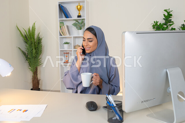 استخدام جهاز تقني حديث ومتطور، سيدة سعودية تجلس خلف الطاولة وتحتسي المشروب المفضل في المكتب، اجراء مكالمة هاتفية عبر جهاز الجوال، تبادل الأحاديث والأخبار والتواصل مع الأهل والأصدقاء،  امراة اماراتية ترتدي العباءة والحجاب تمسك بيدها الهاتف المحمول وتشرب القهوة
