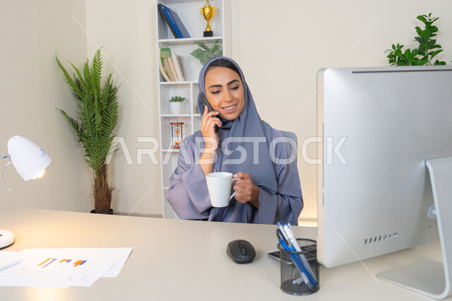 Making a phone call on a mobile device, communicating and exchanging conversations and news with family and friends, an Emirati woman wearing an abaya and hijab holding a mobile phone and drinking coffee, using a modern and advanced technical device, a Saudi woman sitting behind a table and drinking her favorite drink in the office