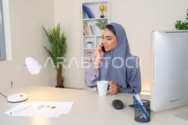 تبادل الأحاديث والأخبار والتواصل مع الأهل والأصدقاء،  امراة اماراتية ترتدي العباءة والحجاب تمسك بيدها الهاتف المحمول وتشرب القهوة، استخدام جهاز تقني حديث ومتطور، سيدة سعودية تجلس خلف الطاولة وتحتسي المشروب المفضل في المكتب، اجراء مكالمة هاتفية عبر جهاز الجوال