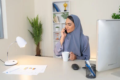 تبادل الأحاديث والأخبار والتواصل مع الأهل والأصدقاء،  امراة اماراتية ترتدي العباءة والحجاب تمسك بيدها الهاتف المحمول وتشرب القهوة، استخدام جهاز تقني حديث ومتطور، سيدة سعودية تجلس خلف الطاولة وتحتسي المشروب المفضل في المكتب، اجراء مكالمة هاتفية عبر جهاز الجوال
