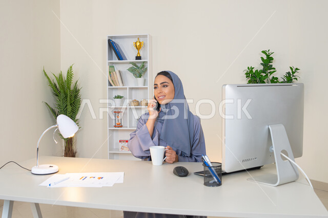 التواصل وتبادل الأحاديث والأخبار مع الأهل والأصدقاء،  امراة اماراتية ترتدي العباءة والحجاب تمسك بيدها الهاتف المحمول وتشرب القهوة، استخدام جهاز تقني حديث ومتطور، سيدة سعودية تجلس خلف الطاولة وتحتسي المشروب المفضل في المكتب، اجراء مكالمة هاتفية عبر جهاز الجوال