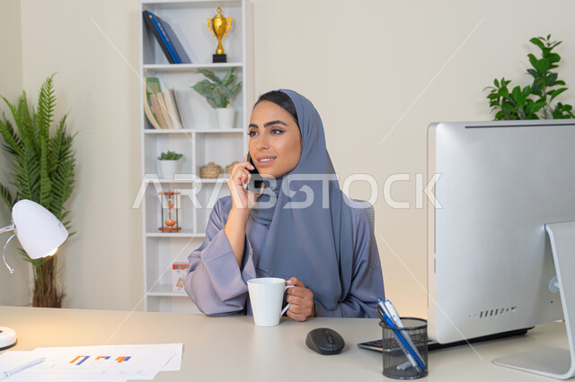 Making a phone call on a mobile device, communicating and exchanging conversations and news with family and friends, an Emirati woman wearing an abaya and hijab holding a mobile phone and drinking coffee, using a modern and advanced technical device, a Saudi woman sitting behind a table and drinking her favorite drink in the office