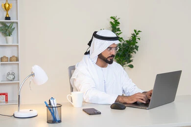 تطور الشركات الاماراتية، إتمام مهام العمل في المكتب، إدارة شؤون الأعمال وتنظيمها، احتساء مشروب القهوة، استخدام جهاز تقني حديث ومتطور، رجل عربي خليجي اماراتي يرتدي الكندورة والغترة البيضاء يستخدم الحاسوب، مهنة ووظيفة مكتبية، دمج العمل مع التقنية والتكنولوجيا