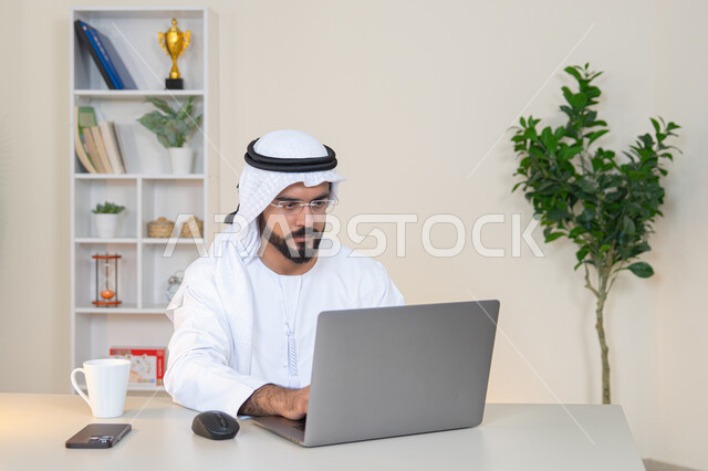 دمج العمل مع التقنية والتكنولوجيا، تطور الشركات الاماراتية، إتمام مهام العمل في المكتب، إدارة شؤون الأعمال وتنظيمها، احتساء مشروب القهوة، استخدام جهاز تقني حديث ومتطور، رجل عربي خليجي اماراتي يرتدي الكندورة والغترة البيضاء يستخدم الحاسوب، مهنة ووظيفة مكتبية