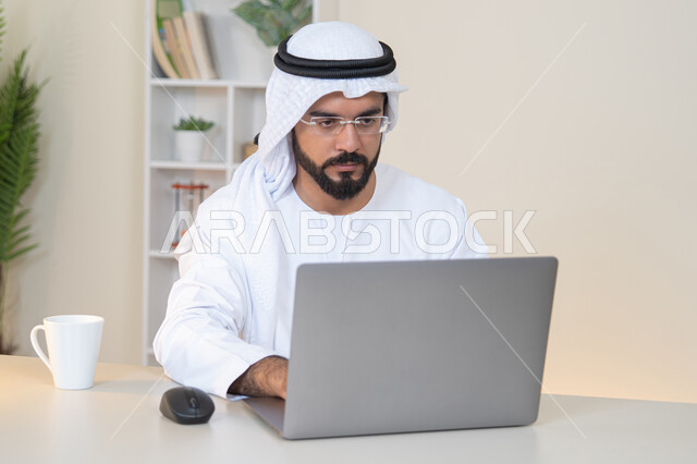 تطور الشركات الاماراتية، إتمام مهام العمل في المكتب، إدارة شؤون الأعمال وتنظيمها، احتساء مشروب القهوة، استخدام جهاز تقني حديث ومتطور، رجل عربي خليجي اماراتي يرتدي الكندورة والغترة البيضاء يستخدم الحاسوب، مهنة ووظيفة مكتبية، دمج العمل مع التقنية والتكنولوجيا