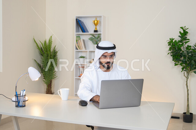 دمج العمل مع التقنية والتكنولوجيا، تطور الشركات الاماراتية، إتمام مهام العمل في المكتب، إدارة شؤون الأعمال وتنظيمها، احتساء مشروب القهوة، استخدام جهاز تقني حديث ومتطور، رجل عربي خليجي اماراتي يرتدي الكندورة والغترة البيضاء يستخدم الحاسوب، مهنة ووظيفة مكتبية