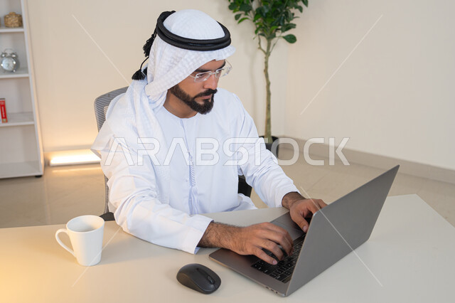 إدارة شؤون الأعمال وتنظيمها، احتساء مشروب القهوة، استخدام جهاز تقني حديث ومتطور، رجل عربي خليجي اماراتي يرتدي الكندورة والغترة البيضاء يستخدم الحاسوب، مهنة ووظيفة مكتبية، دمج العمل مع التقنية والتكنولوجيا، تطور الشركات الاماراتية، إتمام مهام العمل في المكتب