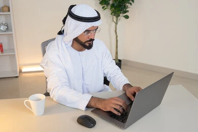إدارة شؤون الأعمال وتنظيمها، احتساء مشروب القهوة، استخدام جهاز تقني حديث ومتطور، رجل عربي خليجي اماراتي يرتدي الكندورة والغترة البيضاء يستخدم الحاسوب، مهنة ووظيفة مكتبية، دمج العمل مع التقنية والتكنولوجيا، تطور الشركات الاماراتية، إتمام مهام العمل في المكتب