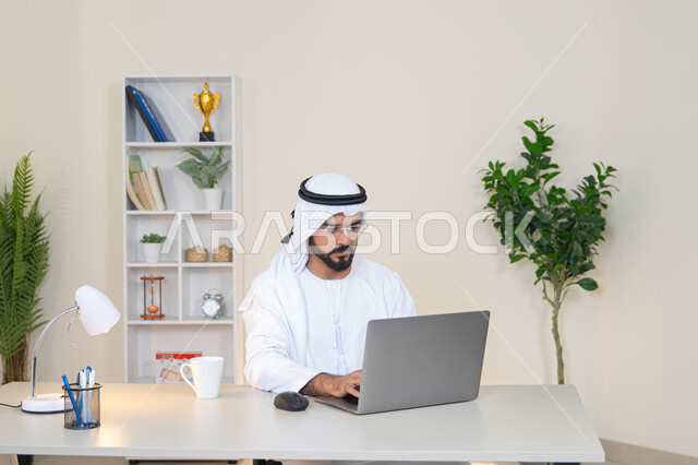 دمج العمل مع التقنية والتكنولوجيا، تطور الشركات الاماراتية، إتمام مهام العمل في المكتب، إدارة شؤون الأعمال وتنظيمها، احتساء مشروب القهوة، استخدام جهاز تقني حديث ومتطور، رجل عربي خليجي اماراتي يرتدي الكندورة والغترة البيضاء يستخدم الحاسوب، مهنة ووظيفة مكتبية